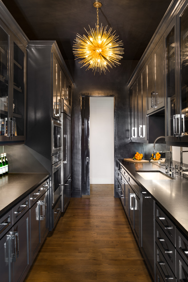 Photo of a contemporary galley kitchen in Dallas with an undermount sink, recessed-panel cabinets, black cabinets, stainless steel appliances, dark hardwood floors, no island, brown floor, black benchtop and mirror splashback.