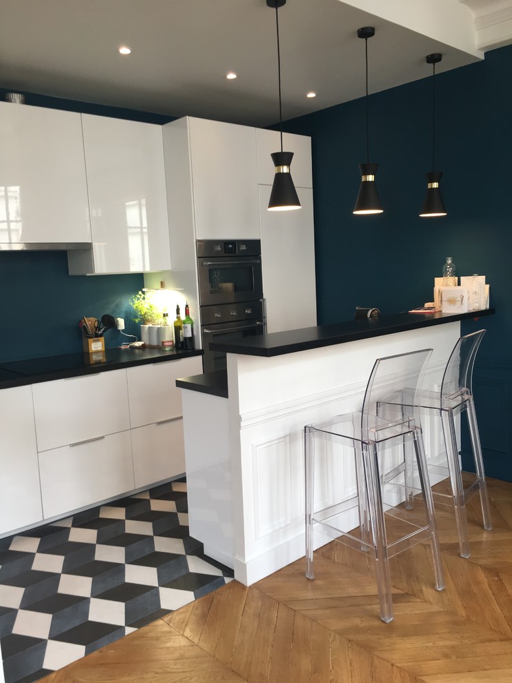 Photo of a mid-sized transitional galley open plan kitchen in Paris with flat-panel cabinets, white cabinets, laminate benchtops, blue splashback, stainless steel appliances, cement tiles, a peninsula, blue floor and black benchtop.