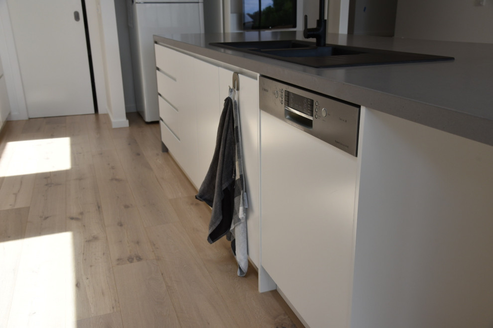 Example of a large danish galley light wood floor, beige floor and tray ceiling open concept kitchen design in Melbourne with a double-bowl sink, flat-panel cabinets, white cabinets, solid surface countertops, white backsplash, glass sheet backsplash, stainless steel appliances, an island and gray countertops