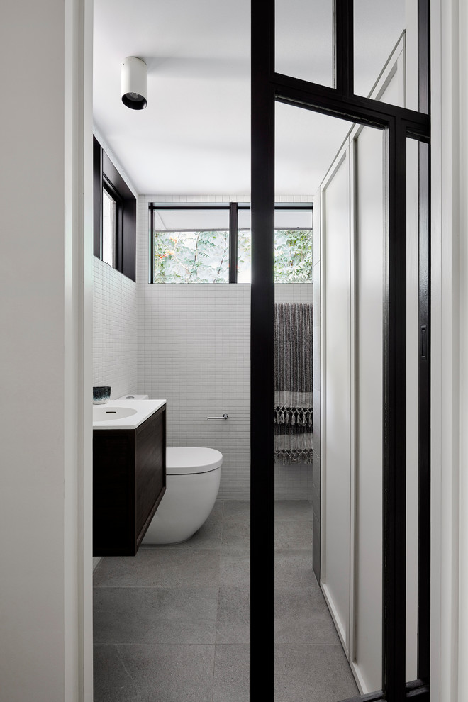 Small traditional grey and brown ensuite half tiled bathroom in Melbourne with freestanding cabinets, black cabinets, an alcove shower, a wall mounted toilet, white tiles, ceramic tiles, white walls, cement flooring, an integrated sink, solid surface worktops, grey floors, an open shower, white worktops, a single sink and a floating vanity unit.
