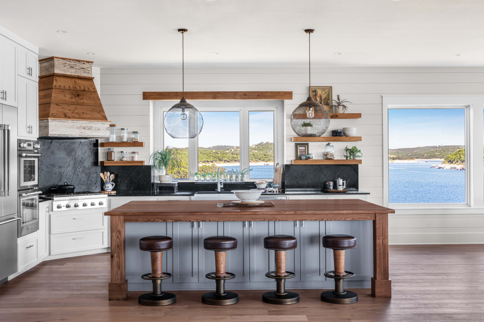 Design ideas for a large beach style l-shaped eat-in kitchen in Austin with a farmhouse sink, shaker cabinets, grey cabinets, black splashback, stone slab splashback, stainless steel appliances, medium hardwood floors, with island, brown floor, black benchtop and wood benchtops.