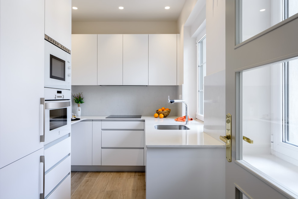 Photo of a contemporary u-shaped separate kitchen in Bilbao with flat-panel cabinets, white cabinets, white splashback, white benchtop, quartz benchtops, white appliances, an undermount sink, light hardwood floors and beige floor.