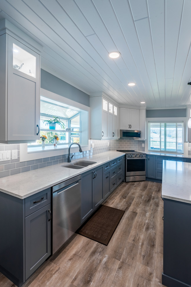 This is an example of a mid-sized traditional l-shaped open plan kitchen in Vancouver with an undermount sink, shaker cabinets, blue cabinets, quartz benchtops, blue splashback, subway tile splashback, stainless steel appliances, vinyl floors, with island, brown floor and white benchtop.