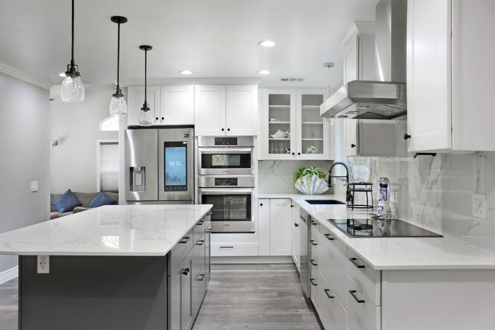 Modern inredning av ett avskilt, stort vit vitt l-kök, med en undermonterad diskho, skåp i shakerstil, vita skåp, bänkskiva i kvarts, vitt stänkskydd, stänkskydd i sten, rostfria vitvaror, mellanmörkt trägolv, en köksö och grått golv