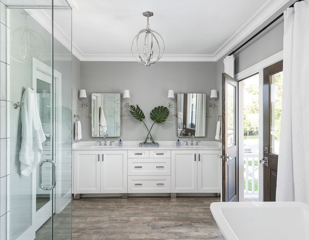 Inspiration for a large transitional master bathroom in Chicago with white cabinets, a freestanding tub, gray tile, grey walls, porcelain floors, an undermount sink, engineered quartz benchtops, brown floor, white benchtops and recessed-panel cabinets.