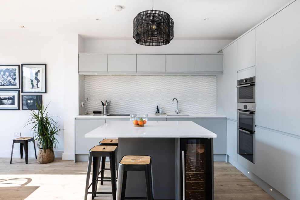 This is an example of a contemporary l-shaped kitchen in Hertfordshire with an undermount sink, flat-panel cabinets, grey cabinets, white splashback, mosaic tile splashback, light hardwood floors, with island and white benchtop.