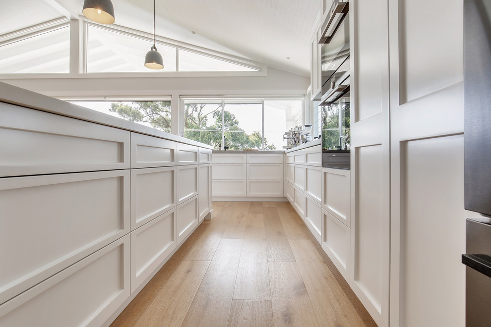 Mid-sized beach style l-shaped open plan kitchen in Sydney with a double-bowl sink, shaker cabinets, white cabinets, quartz benchtops, grey splashback, stainless steel appliances, medium hardwood floors, with island, brown floor and white benchtop.