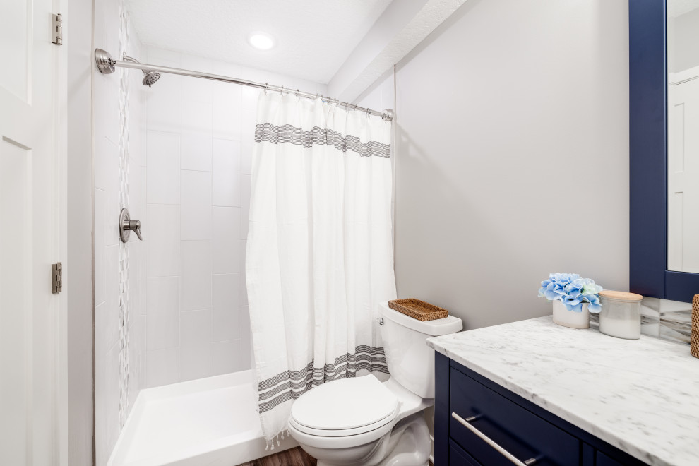 Klassisk inredning av ett mellanstort vit vitt badrum med dusch, med skåp i shakerstil, blå skåp, en dusch i en alkov, en toalettstol med hel cisternkåpa, vit kakel, porslinskakel, grå väggar, vinylgolv, ett undermonterad handfat, bänkskiva i kvarts, brunt golv och dusch med duschdraperi
