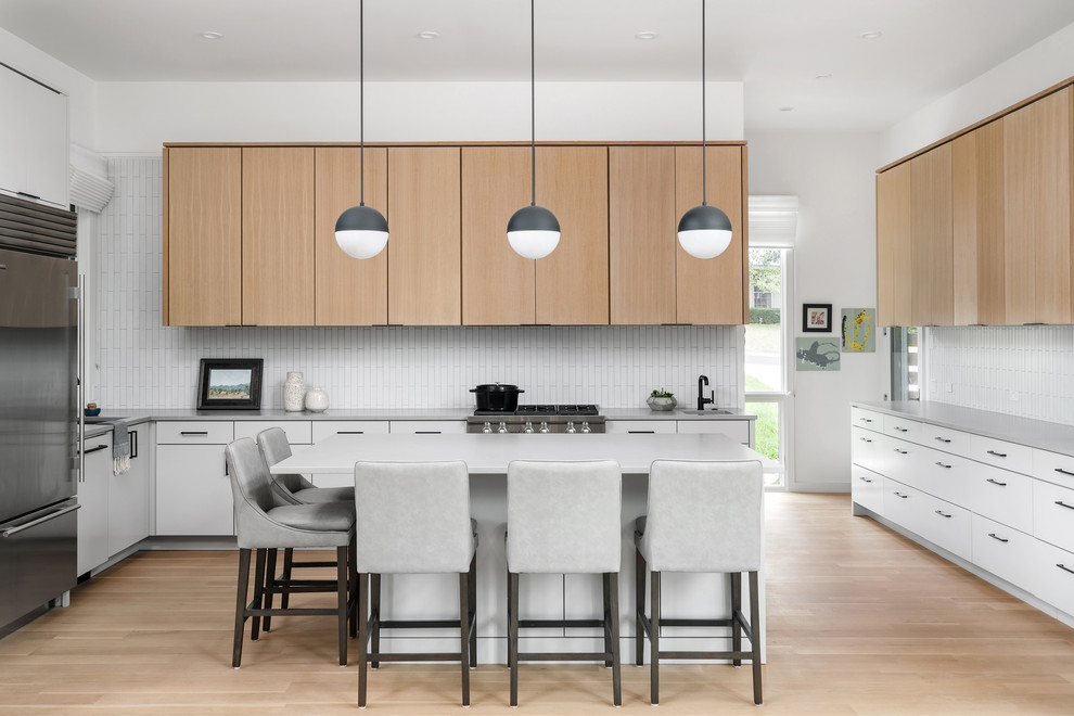 Photo of a contemporary u-shaped kitchen in Austin with flat-panel cabinets, light wood cabinets, white splashback, stainless steel appliances, light hardwood floors, with island, beige floor and grey benchtop.