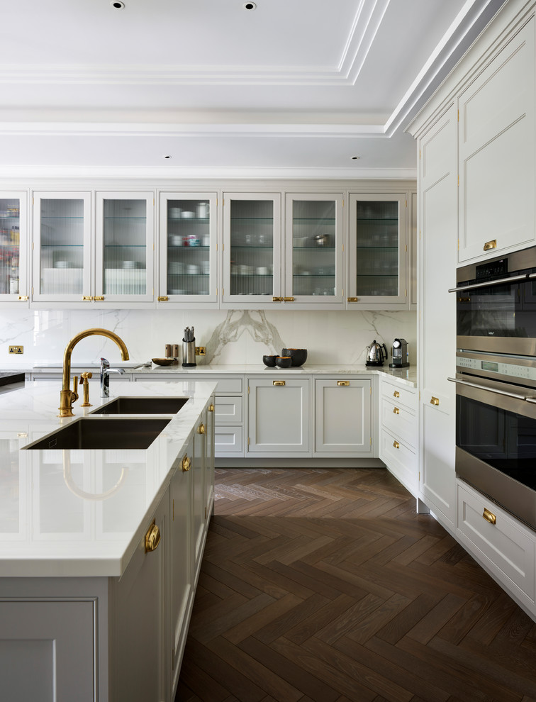 This is an example of a large transitional l-shaped eat-in kitchen in London with with island, a double-bowl sink, beaded inset cabinets, beige cabinets, white splashback, stone slab splashback, stainless steel appliances, dark hardwood floors and brown floor.