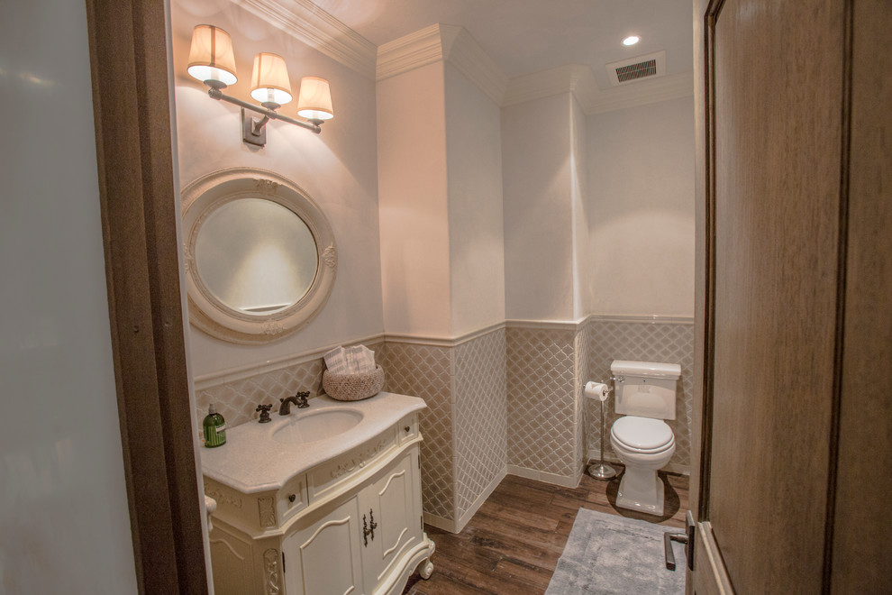 Photo of a small traditional powder room in Santa Barbara with white walls, an undermount sink, white cabinets, beige tile, ceramic tile, medium hardwood floors, granite benchtops, furniture-like cabinets, a two-piece toilet and brown floor.