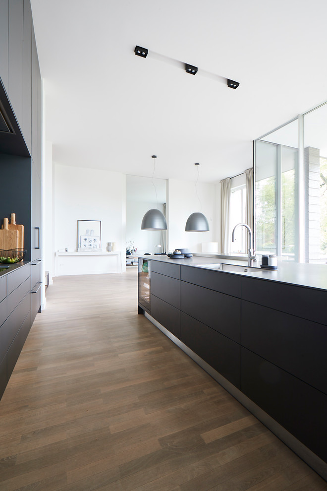 Design ideas for a large contemporary single-wall eat-in kitchen in Hamburg with flat-panel cabinets, black cabinets, black splashback, black appliances, light hardwood floors, with island, beige floor, black benchtop, a single-bowl sink and concrete benchtops.
