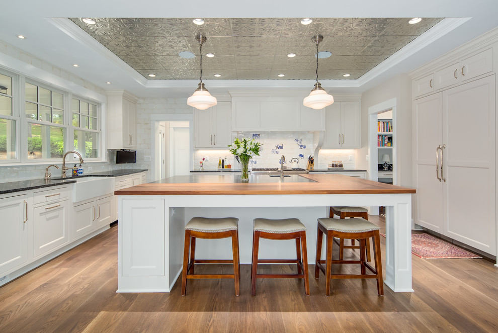 This is an example of a transitional u-shaped kitchen in DC Metro with a farmhouse sink, shaker cabinets, white cabinets, wood benchtops, white splashback, dark hardwood floors, with island, brown floor and brown benchtop.