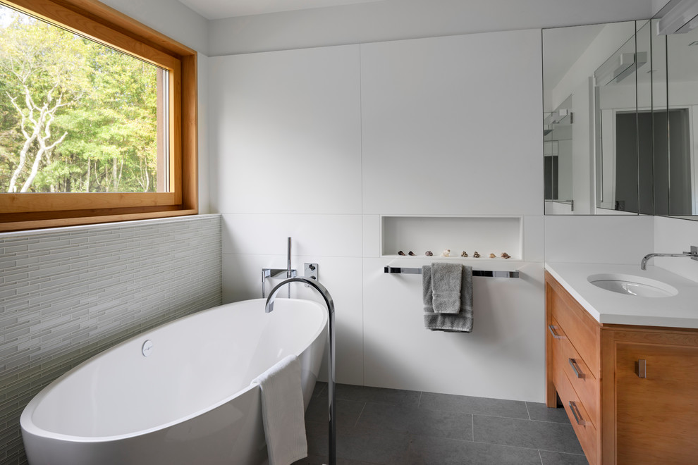 Mid-sized contemporary master bathroom in Providence with medium wood cabinets, a freestanding tub, white tile, glass tile, white walls, porcelain floors, solid surface benchtops, grey floor, white benchtops, an undermount sink and flat-panel cabinets.