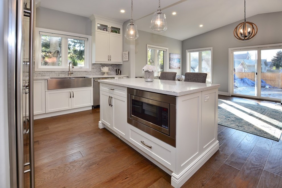 Eat-in kitchen - mid-sized transitional l-shaped medium tone wood floor and brown floor eat-in kitchen idea in Toronto with a farmhouse sink, shaker cabinets, white cabinets, quartz countertops, gray backsplash, marble backsplash, stainless steel appliances, an island and white countertops