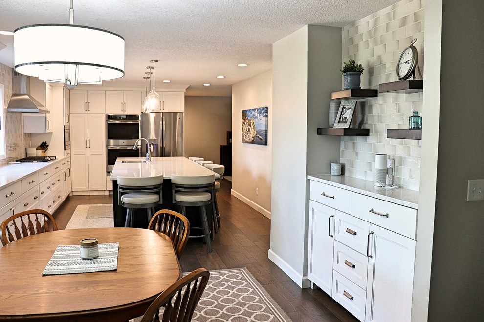 Example of a transitional l-shaped medium tone wood floor and brown floor kitchen pantry design in Cedar Rapids with an undermount sink, recessed-panel cabinets, white cabinets, quartzite countertops, gray backsplash, porcelain backsplash, stainless steel appliances, an island and white countertops