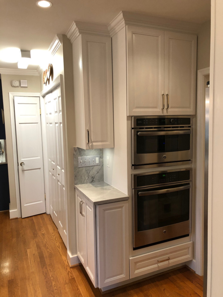 Photo of a medium sized classic u-shaped kitchen/diner in DC Metro with a submerged sink, recessed-panel cabinets, white cabinets, quartz worktops, white splashback, engineered quartz splashback, stainless steel appliances, medium hardwood flooring, an island, brown floors and white worktops.
