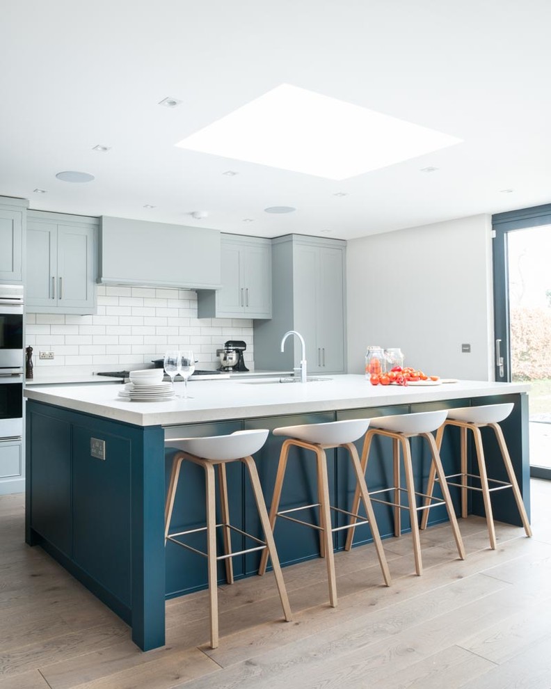 This is an example of a large contemporary single-wall open plan kitchen in Kent with recessed-panel cabinets, blue cabinets, solid surface benchtops, white splashback, ceramic splashback, stainless steel appliances, light hardwood floors and with island.
