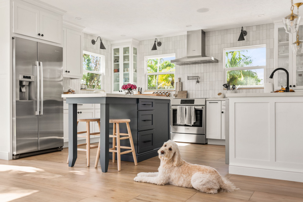 Inspiration for a mid-sized beach style u-shaped eat-in kitchen in Tampa with an undermount sink, shaker cabinets, white cabinets, quartz benchtops, white splashback, porcelain splashback, stainless steel appliances, medium hardwood floors, with island, brown floor and white benchtop.