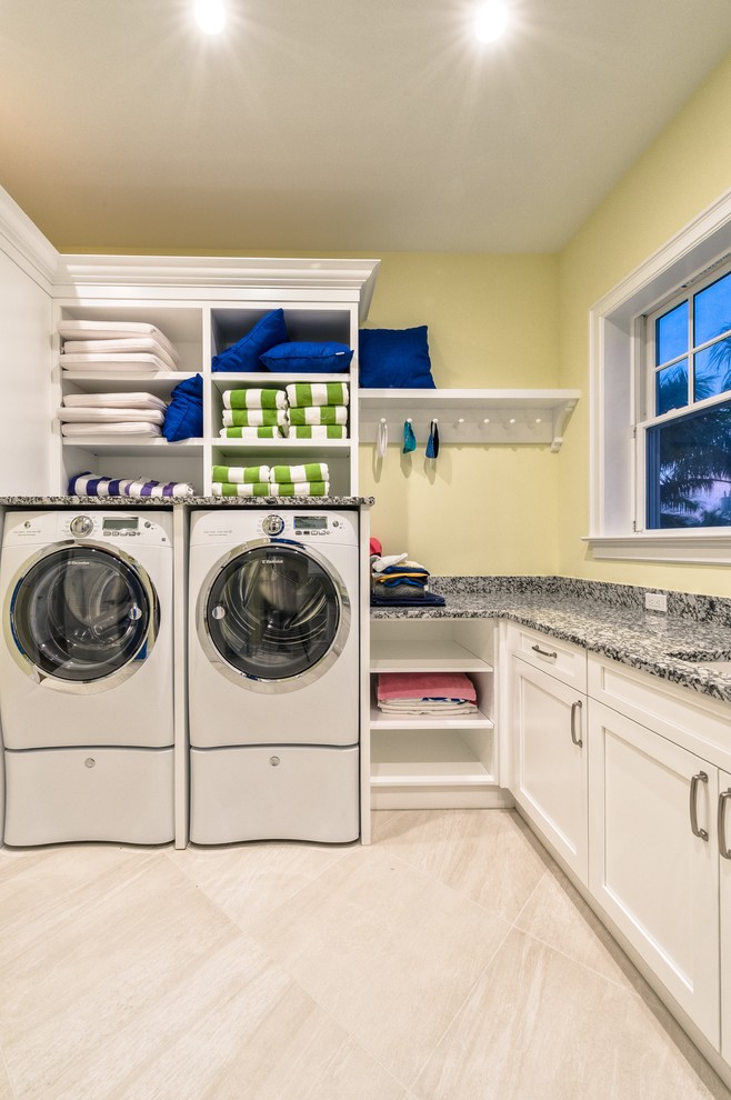 Inspiration for an expansive beach style l-shaped dedicated laundry room in Miami with an undermount sink, recessed-panel cabinets, white cabinets, granite benchtops, yellow walls, porcelain floors and a side-by-side washer and dryer.