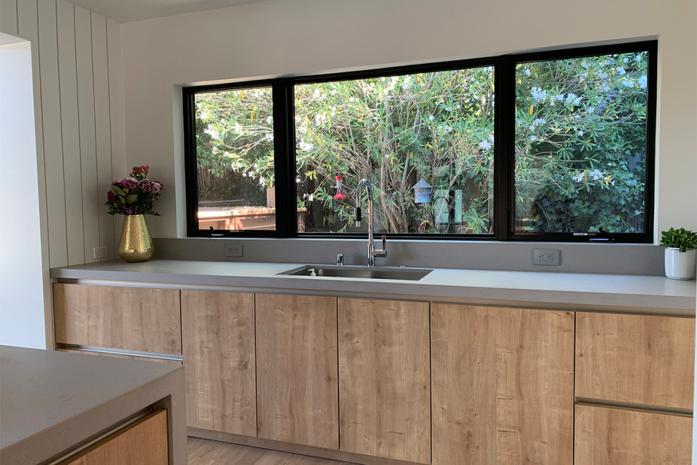 Photo of a medium sized scandinavian l-shaped kitchen/diner in San Francisco with a submerged sink, flat-panel cabinets, black cabinets, composite countertops, grey splashback, stone slab splashback, integrated appliances, light hardwood flooring, an island, brown floors and grey worktops.