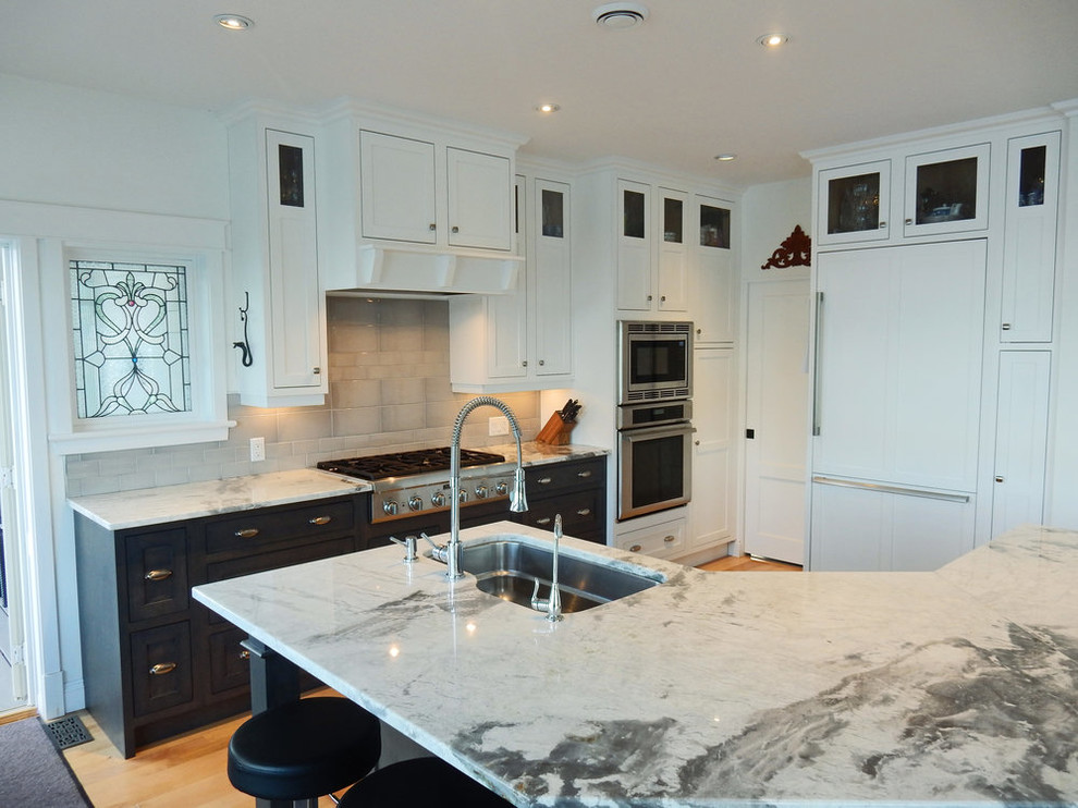 Example of a transitional medium tone wood floor and brown floor kitchen design with an undermount sink, white cabinets, quartz countertops, gray backsplash, ceramic backsplash, stainless steel appliances and white countertops