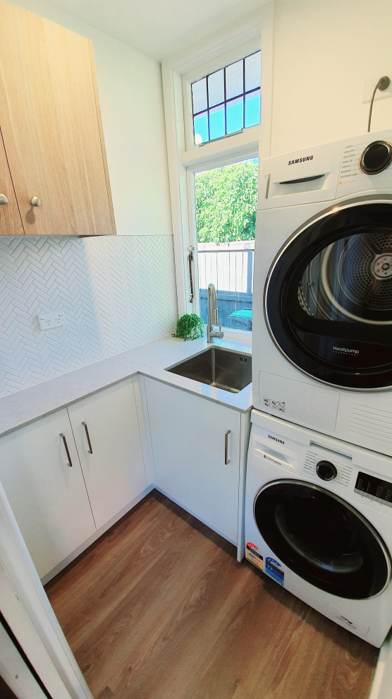 Photo of a small contemporary l-shaped dedicated laundry room in Christchurch with an undermount sink, white cabinets, quartz benchtops, white splashback, porcelain splashback, white walls, laminate floors, a stacked washer and dryer, brown floor and grey benchtop.