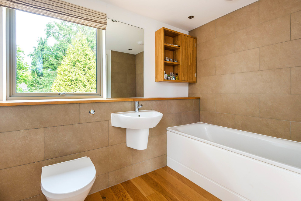 Photo of a large contemporary bathroom in Kent with an alcove tub, a wall-mount toilet, brown tile, brown walls, medium hardwood floors and a wall-mount sink.
