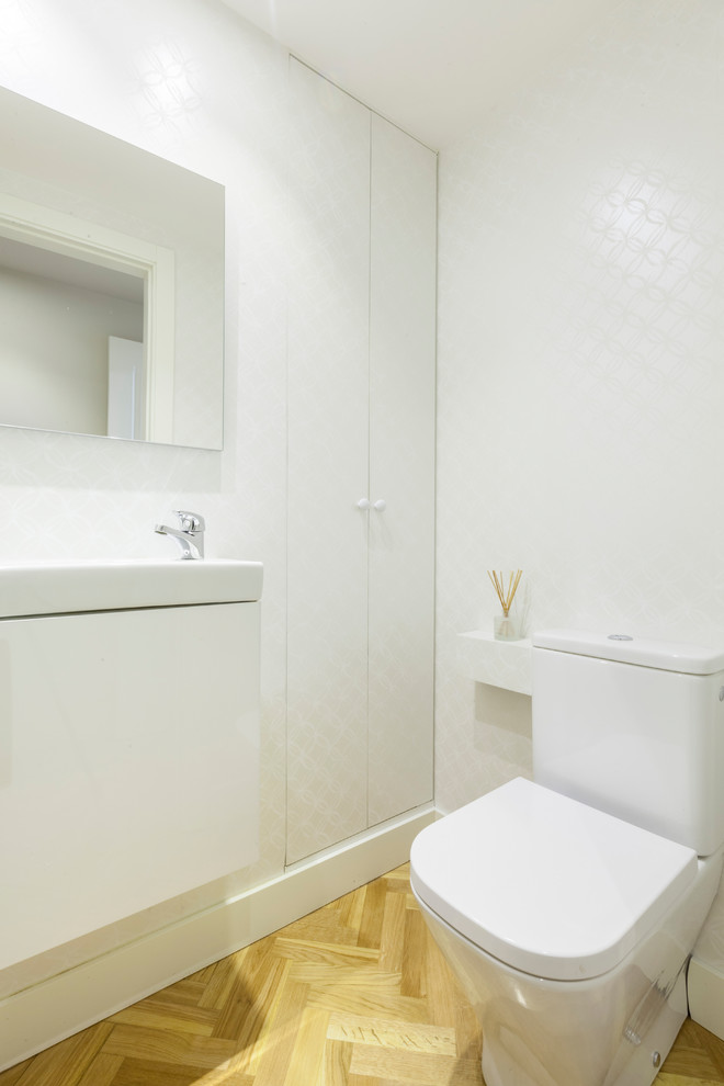 Inspiration for a small transitional powder room in Madrid with flat-panel cabinets, white cabinets, a two-piece toilet, white walls, medium hardwood floors, an integrated sink and brown floor.