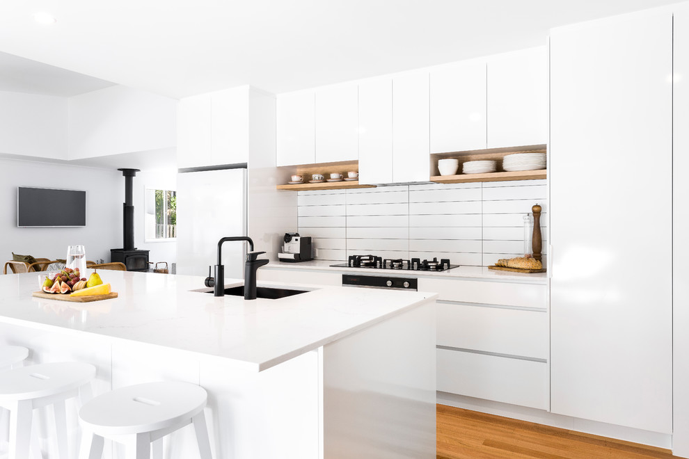 Mid-sized modern galley open plan kitchen in Gold Coast - Tweed with a drop-in sink, flat-panel cabinets, white cabinets, quartz benchtops, white splashback, ceramic splashback, white appliances, medium hardwood floors, with island and white benchtop.