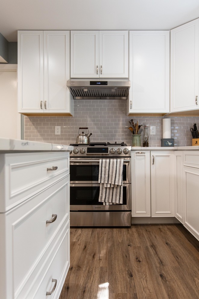 This is an example of a large contemporary open plan kitchen in DC Metro with an undermount sink, raised-panel cabinets, white cabinets, quartz benchtops, grey splashback, subway tile splashback, stainless steel appliances, vinyl floors, with island, brown floor and white benchtop.
