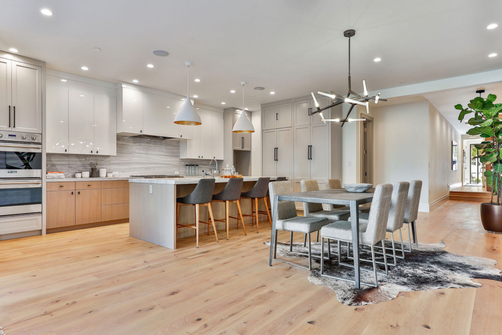 This is an example of a large contemporary l-shaped open plan kitchen in San Francisco with flat-panel cabinets, white cabinets, marble benchtops, marble splashback, stainless steel appliances, light hardwood floors, with island, white benchtop, grey splashback and beige floor.