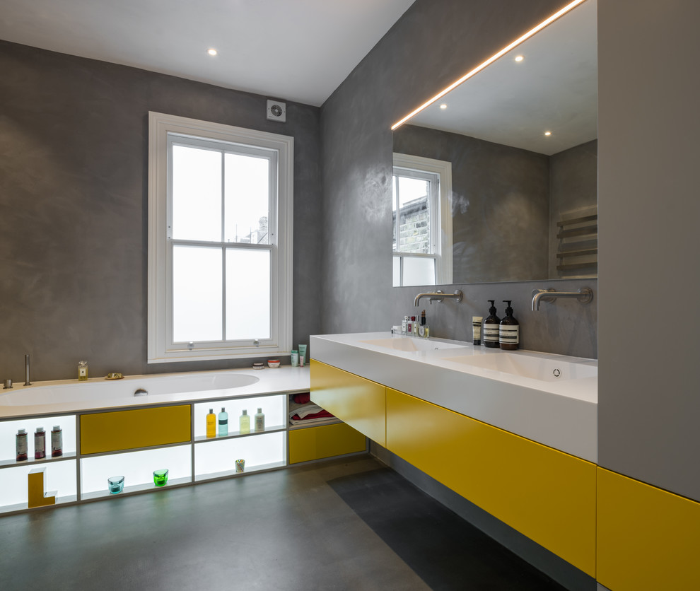 Mid-sized modern bathroom in London with flat-panel cabinets, yellow cabinets, a drop-in tub, grey walls, grey floor, concrete floors, an integrated sink and white benchtops.