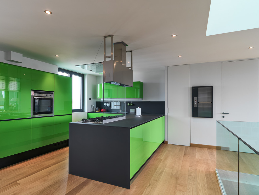 This is an example of a contemporary l-shaped open plan kitchen in Milan with flat-panel cabinets, green cabinets, solid surface benchtops, glass sheet splashback, stainless steel appliances, light hardwood floors and with island.