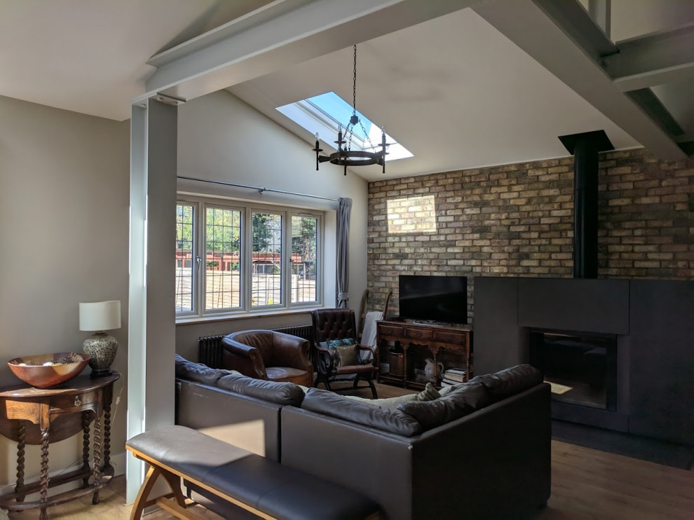 Photo of a mid-sized eclectic living room in London with beige walls, laminate floors, a wood stove, a metal fireplace surround, a freestanding tv, beige floor, exposed beam and brick walls.