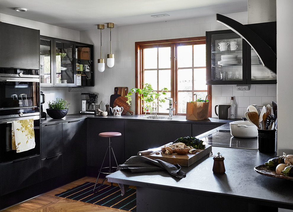 This is an example of a large scandinavian u-shaped open plan kitchen in Other with flat-panel cabinets, black cabinets, white splashback, ceramic splashback, brown floor, an undermount sink, black appliances, medium hardwood floors, no island and black benchtop.