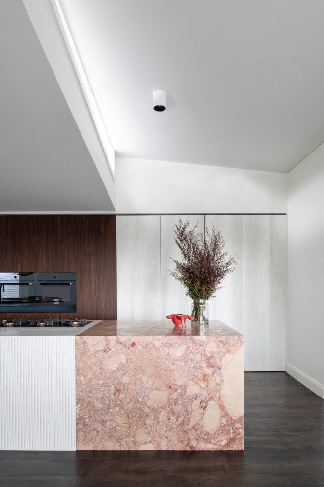 Large contemporary galley open plan kitchen in Sydney with a double-bowl sink, flat-panel cabinets, white cabinets, solid surface benchtops, black appliances, dark hardwood floors, with island and white benchtop.