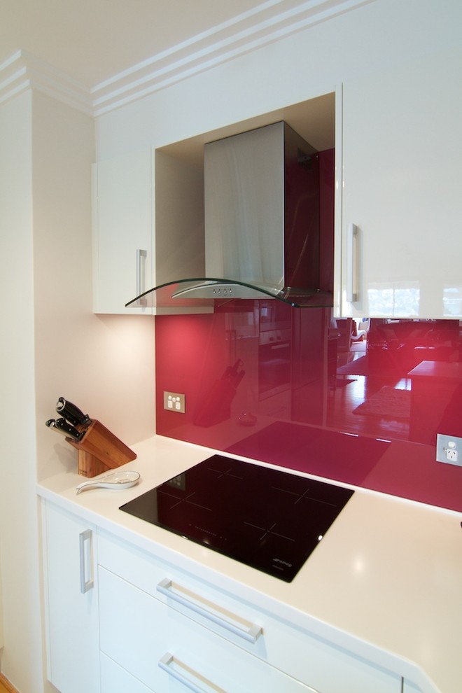 This is an example of a small modern u-shaped separate kitchen in Sydney with an integrated sink, flat-panel cabinets, white cabinets, solid surface benchtops, red splashback, glass sheet splashback, stainless steel appliances, light hardwood floors and no island.