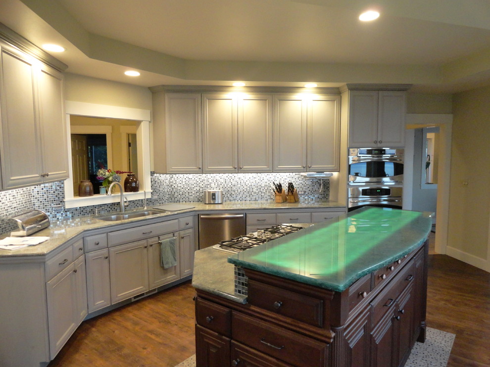 Photo of a large beach style l-shaped eat-in kitchen in Seattle with an undermount sink, distressed cabinets, stainless steel appliances, flat-panel cabinets, green splashback, glass tile splashback, dark hardwood floors, with island and glass benchtops.