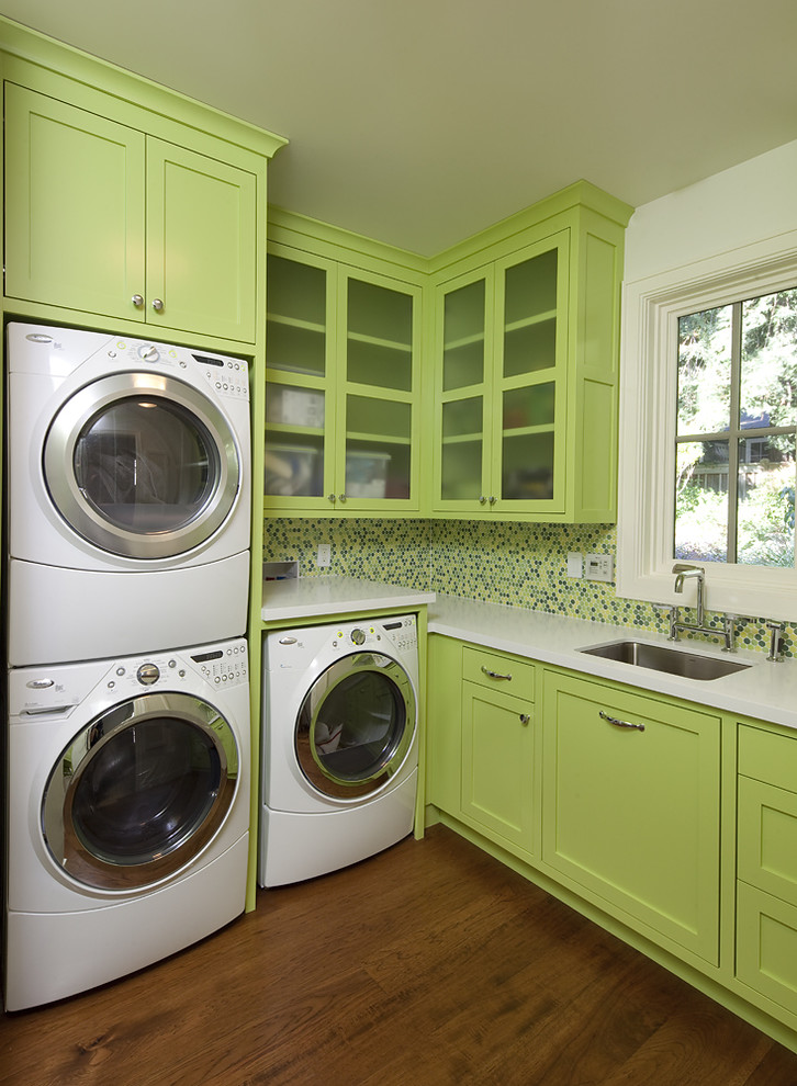 Photo of a contemporary laundry room in San Francisco with green cabinets, solid surface benchtops, a stacked washer and dryer, an undermount sink, brown floor and white benchtop.