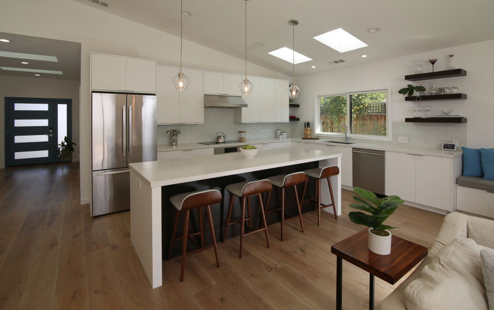 Photo of an expansive contemporary l-shaped eat-in kitchen in San Francisco with a single-bowl sink, flat-panel cabinets, white cabinets, quartz benchtops, white splashback, glass tile splashback, stainless steel appliances, medium hardwood floors, with island, brown floor, vaulted and grey benchtop.