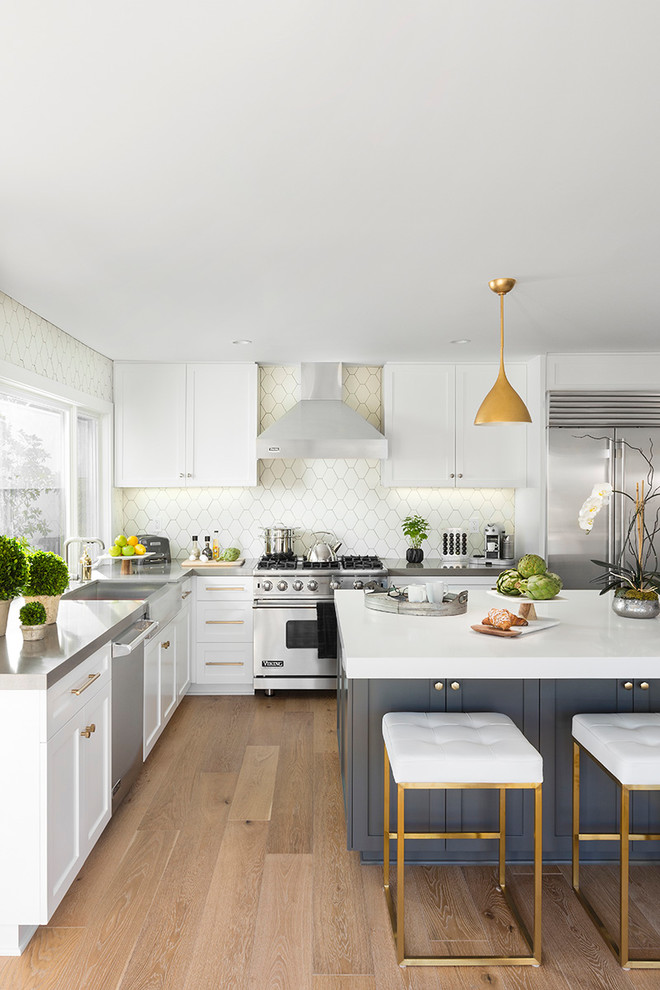 Photo of a mid-sized transitional l-shaped eat-in kitchen in Los Angeles with white cabinets, stainless steel appliances, with island, a farmhouse sink, recessed-panel cabinets, beige splashback, light hardwood floors and beige floor.
