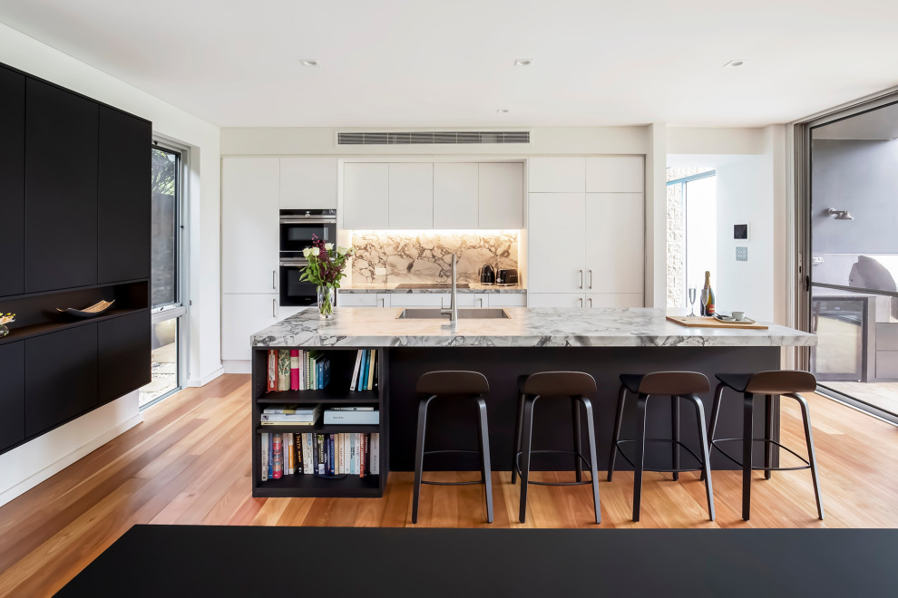 This is an example of a mid-sized contemporary galley open plan kitchen in Sydney with a drop-in sink, flat-panel cabinets, marble benchtops, grey splashback, stone slab splashback, stainless steel appliances, medium hardwood floors, with island and grey benchtop.