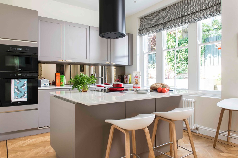 Inspiration for a medium sized traditional enclosed kitchen in London with a built-in sink, raised-panel cabinets, grey cabinets, composite countertops, mirror splashback, black appliances, medium hardwood flooring and an island.