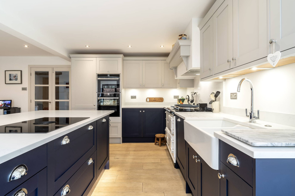 Example of a mid-sized minimalist l-shaped eat-in kitchen design in Oxfordshire with a farmhouse sink, shaker cabinets, white cabinets, quartzite countertops, white backsplash, black appliances, an island and white countertops
