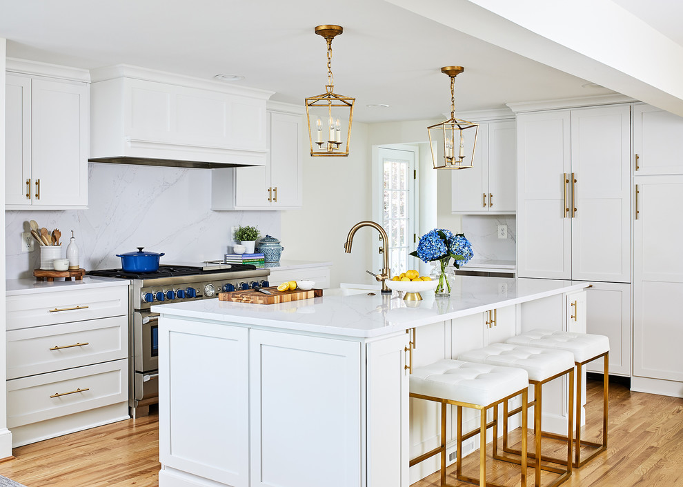 Photo of a transitional kitchen in DC Metro with a farmhouse sink, shaker cabinets, white cabinets, white splashback, stone slab splashback, stainless steel appliances, light hardwood floors, with island and white benchtop.