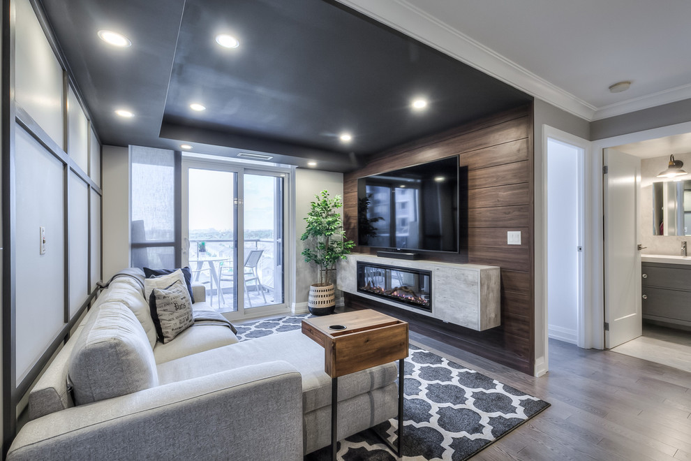 Small contemporary open concept family room in Toronto with grey walls, medium hardwood floors, a hanging fireplace, a wood fireplace surround, a wall-mounted tv and grey floor.