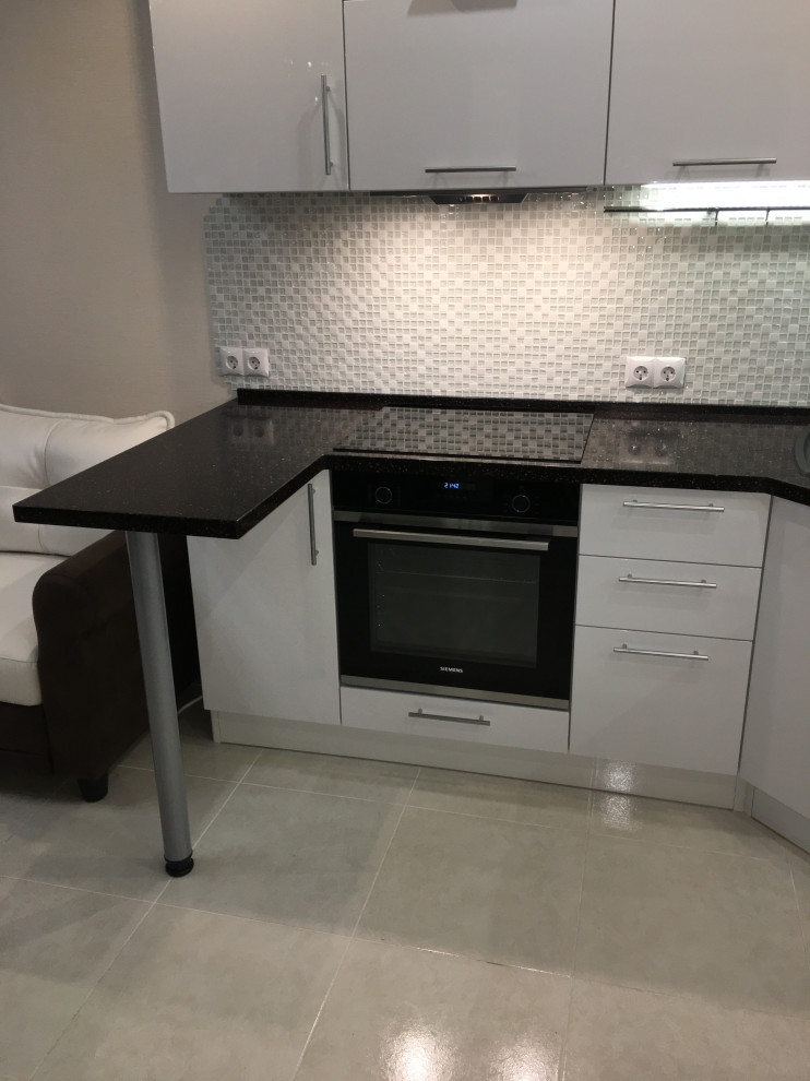 This is an example of a mid-sized contemporary u-shaped open plan kitchen in Other with an undermount sink, flat-panel cabinets, white cabinets, solid surface benchtops, grey splashback, glass tile splashback, stainless steel appliances, ceramic floors, no island, beige floor, black benchtop and coffered.