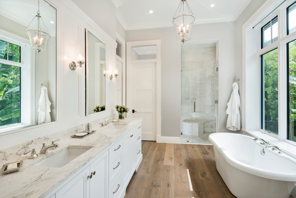 Beach style master bathroom in Miami with recessed-panel cabinets, white cabinets, a freestanding tub, an alcove shower, gray tile, grey walls, medium hardwood floors, an undermount sink, beige floor and a hinged shower door.
