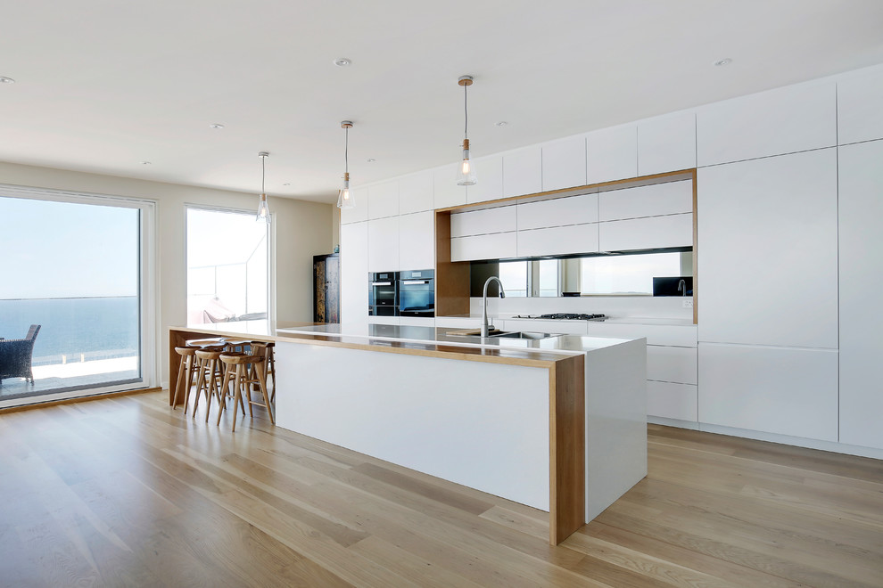 This is an example of a large contemporary galley open plan kitchen in Sydney with a double-bowl sink, quartz benchtops, mirror splashback, black appliances, light hardwood floors, flat-panel cabinets, white cabinets, with island and beige floor.